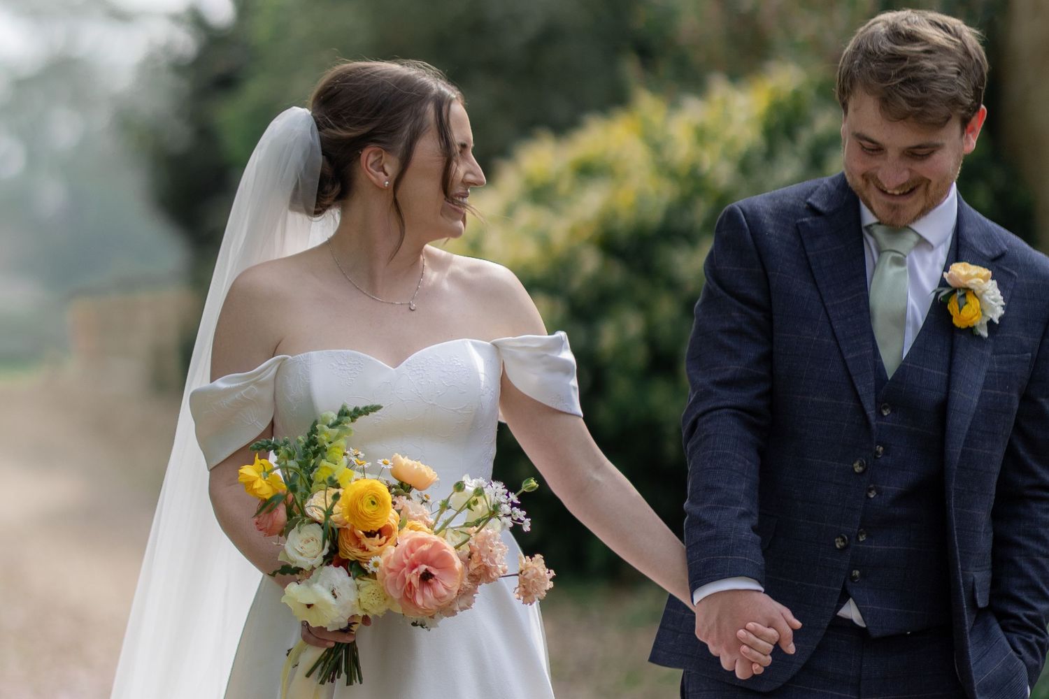 Newlywed couple holding hands outdoors with bride in off-shoulder dress holding colorful bouquet.