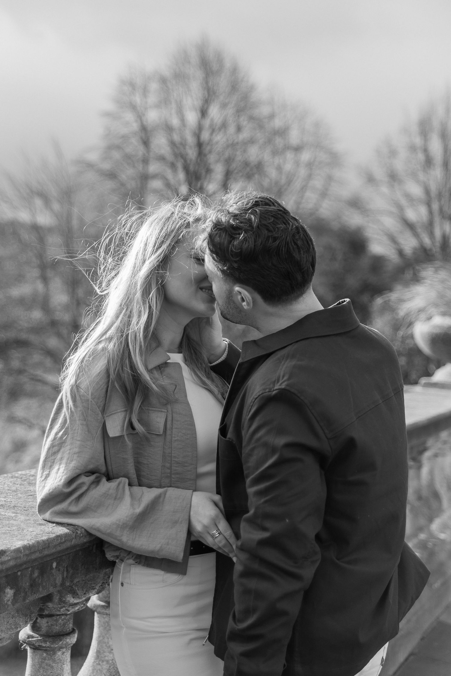 Black and white photo of a couple sharing a romantic kiss outdoors near a stone balustrade.