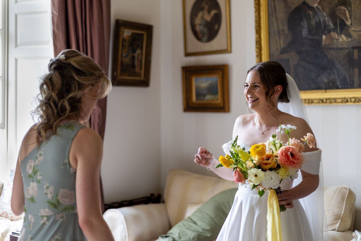 Bride in a flowing white wedding dress gets ready in an elegant room with ornate gold-framed artwork.