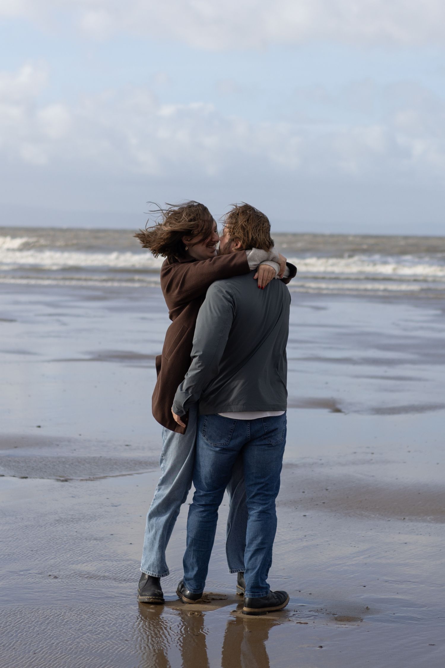 A couple walks together on a windy beach with waves crashing in the background.