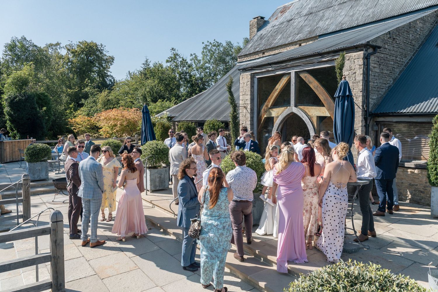 Guests mingle outdoors at a sunny wedding reception venue with stone building and lush greenery.