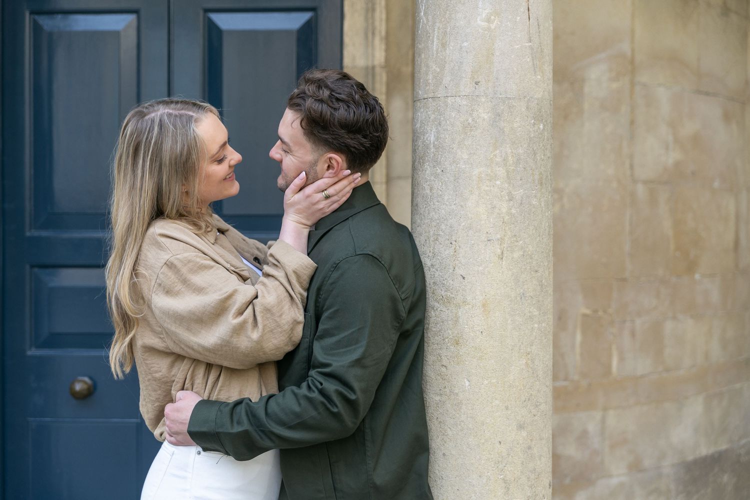 Romantic couple embracing and gazing at each other beside a stone column and dark blue door.