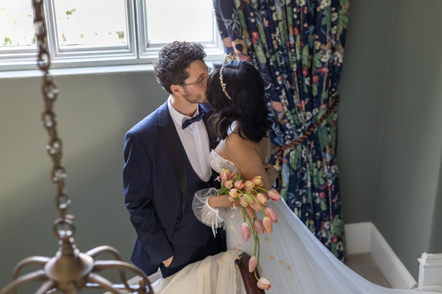 Bride and groom share a romantic kiss near a floral curtain, bride holding a tulip bouquet in a bright room.