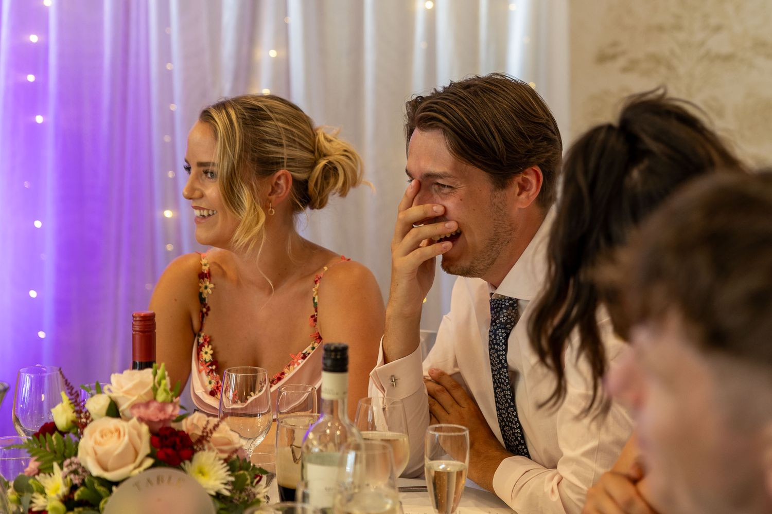 Wedding reception dinner setting with purple lighting and white floral centerpieces.