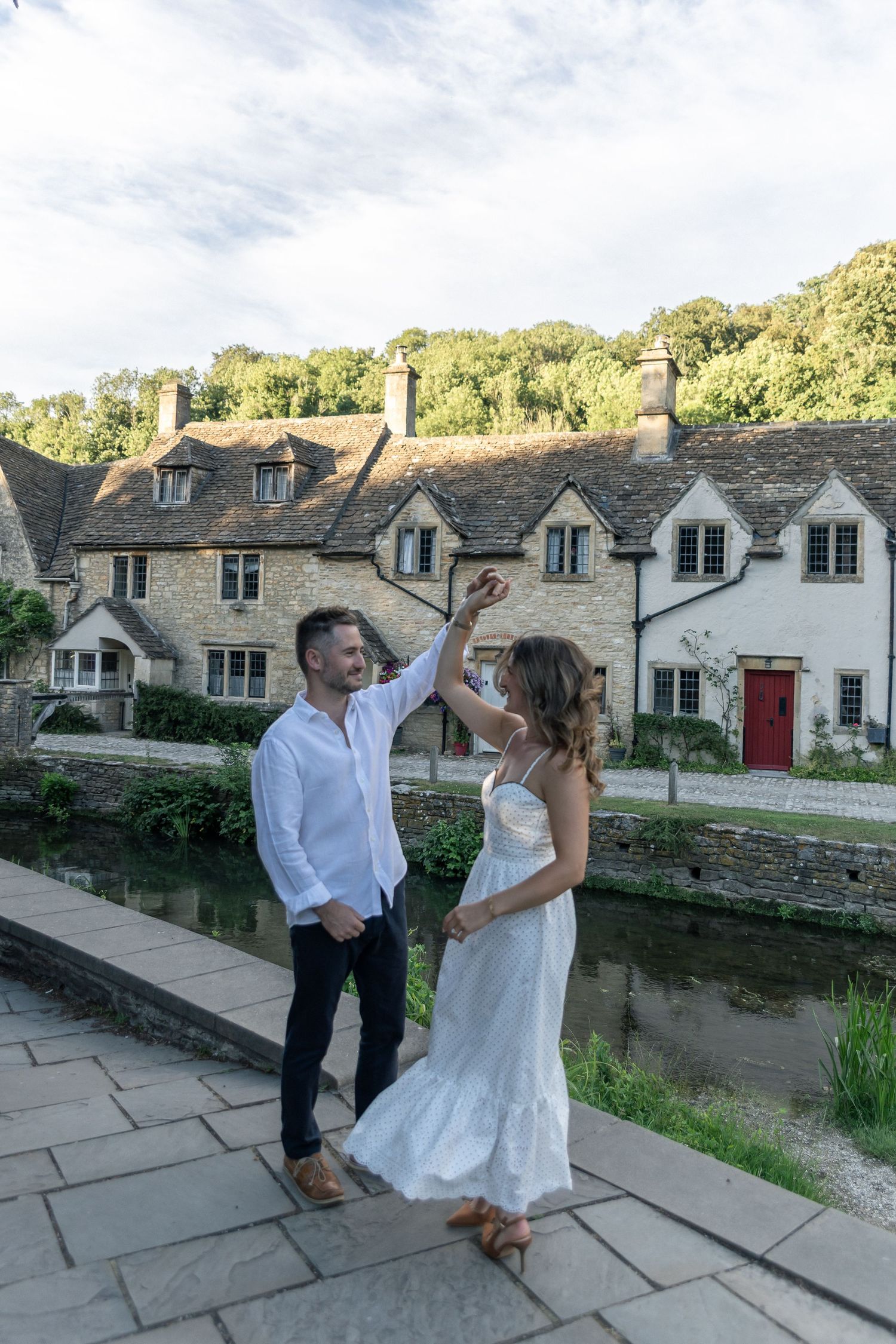 Couple dancing by a river with charming Cotswolds stone cottages in the background on a sunny day.