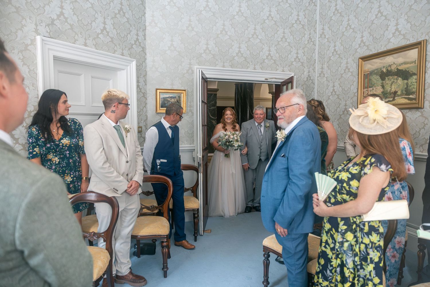 Elegant wedding ceremony in The Guildhall, Bath with guests gathered as bride holds bouquet, groom in beige suit stands nearby.