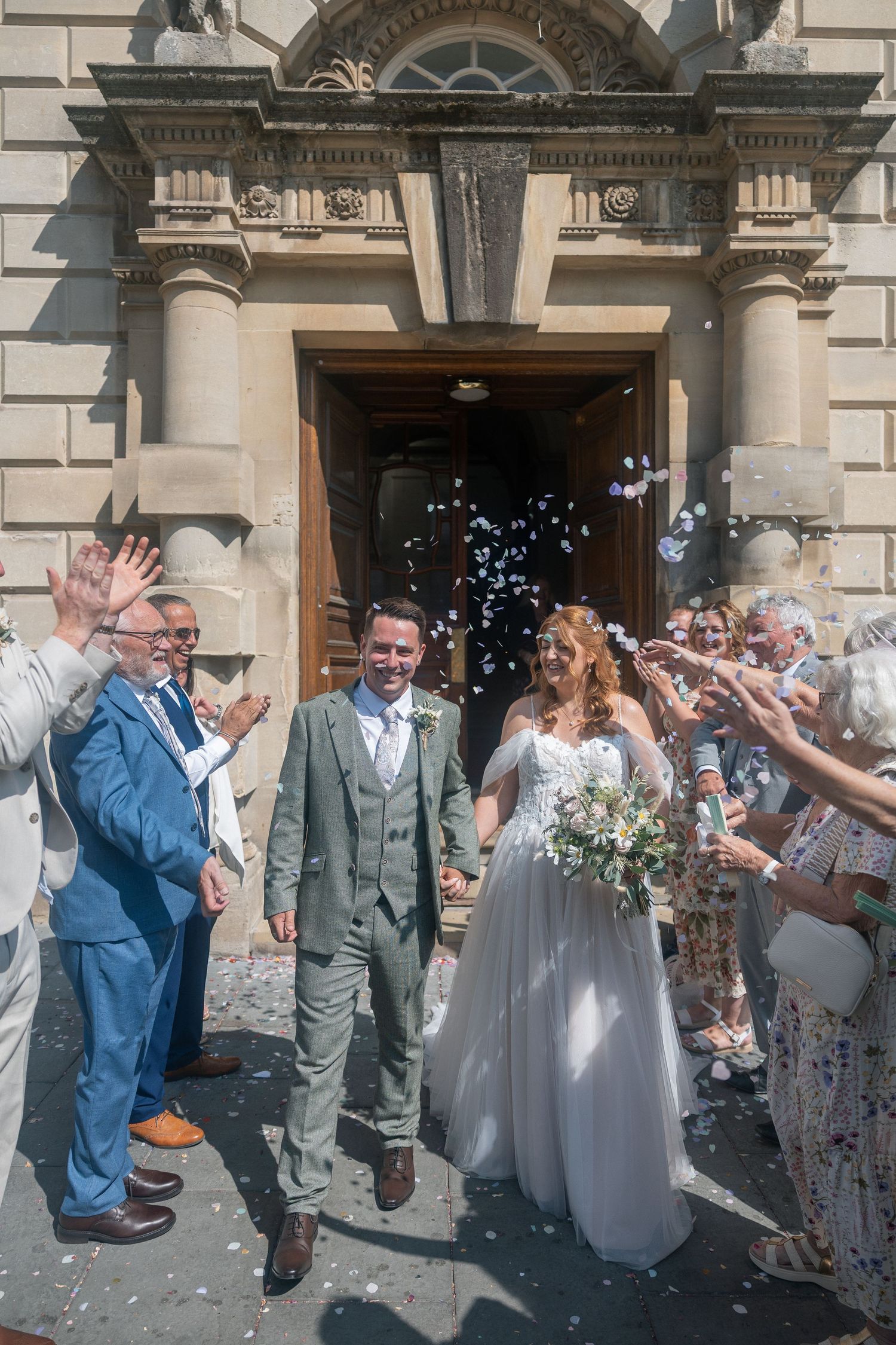 Newlyweds exit The Guildhall Bath as guests throw white confetti in a joyful wedding celebration.