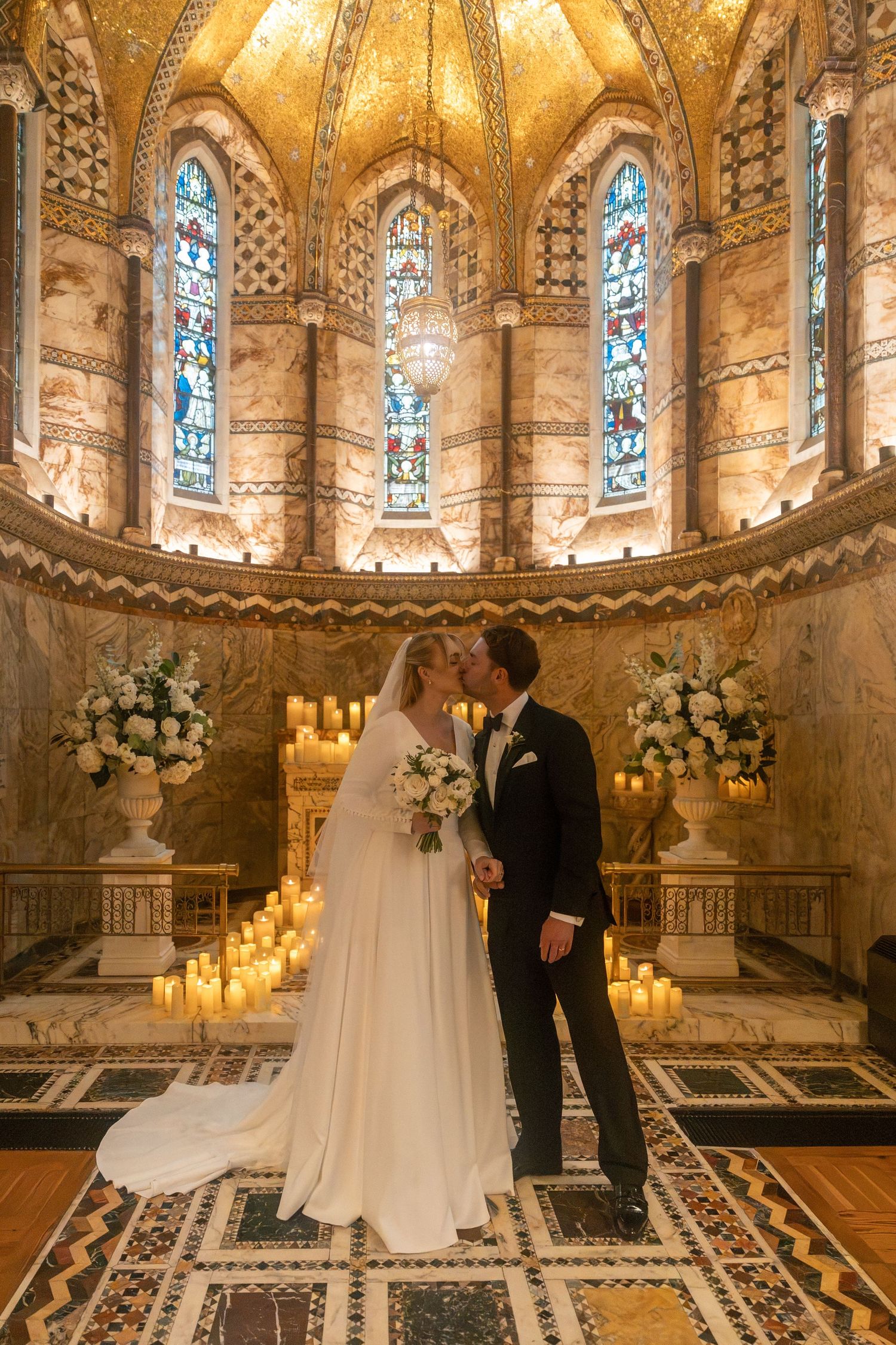 Bride and groom share a kiss in the ornate Fitzrovia chapel with golden mosaics, stained glass windows, and candlelit floral arrangements.