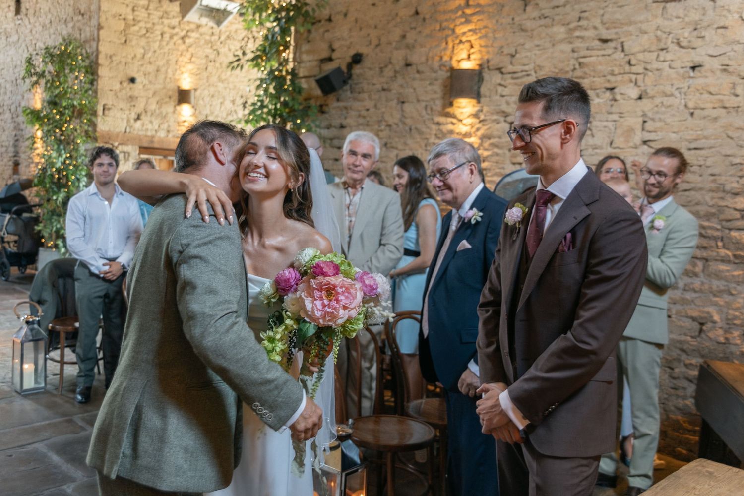 Bride and groom embrace joyfully at rustic stone wedding venue, surrounded by smiling guests holding pink bouquet.
