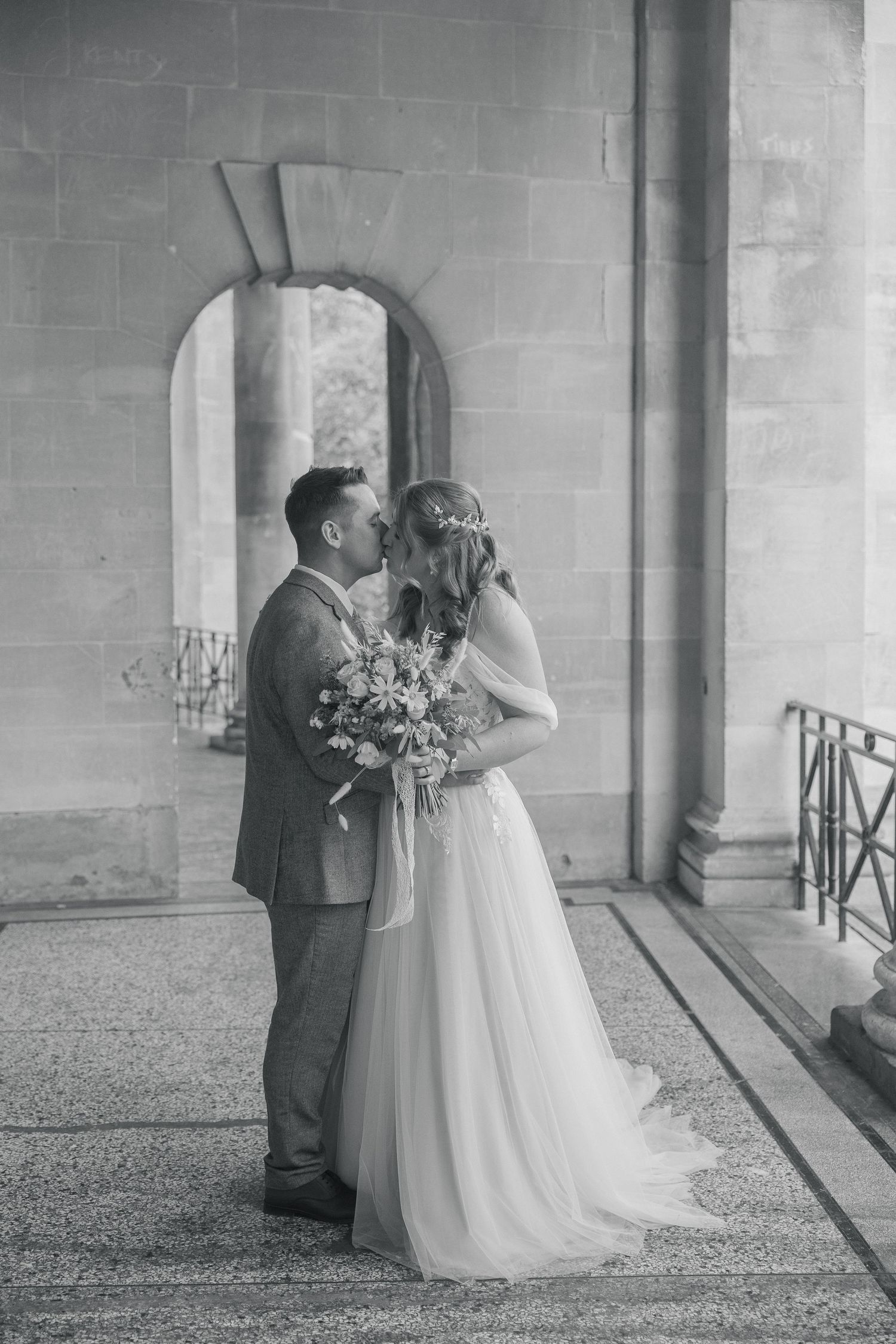 Black and white photo of a bride and groom sharing a kiss, bride holding a bouquet, in Parade Gardens, Bath