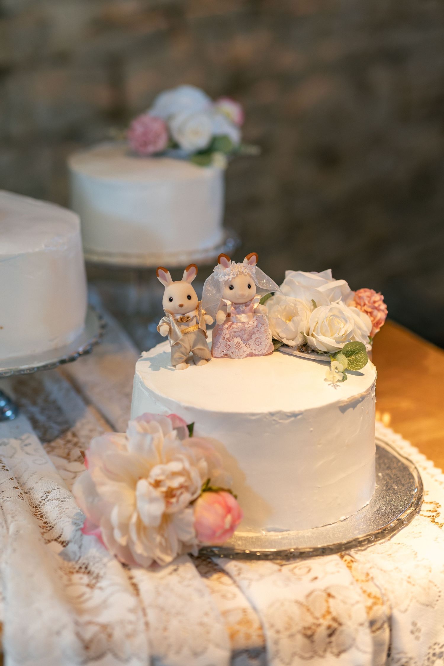 White wedding cake with bunny bride and groom figurines, decorated with pink and white roses on a silver cake stand.