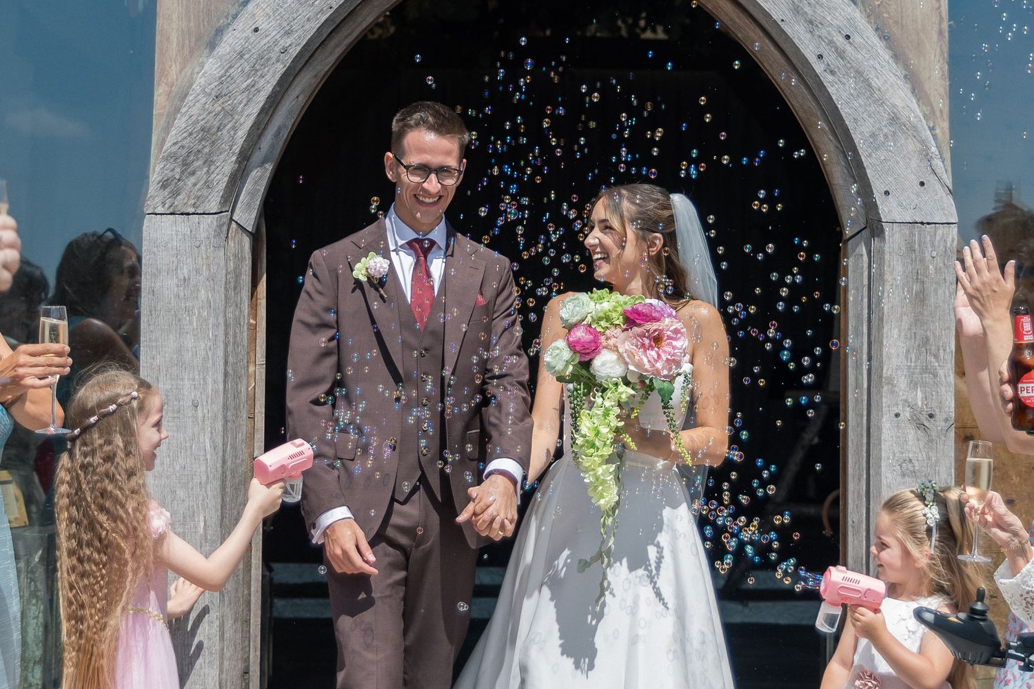 Joyful newlyweds exit church as guests throw colorful confetti, bride holding pink bouquet in white gown, groom in brown suit.