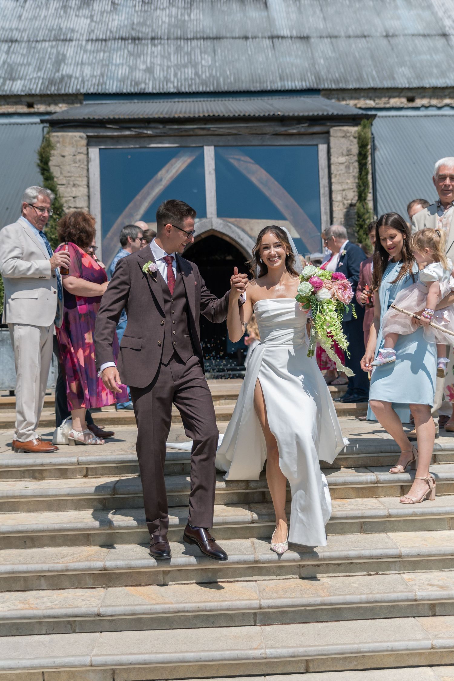 Bride and groom exit Cripps Barn smiling, bride holds pink bouquet in white gown with slit, groom in dark suit.