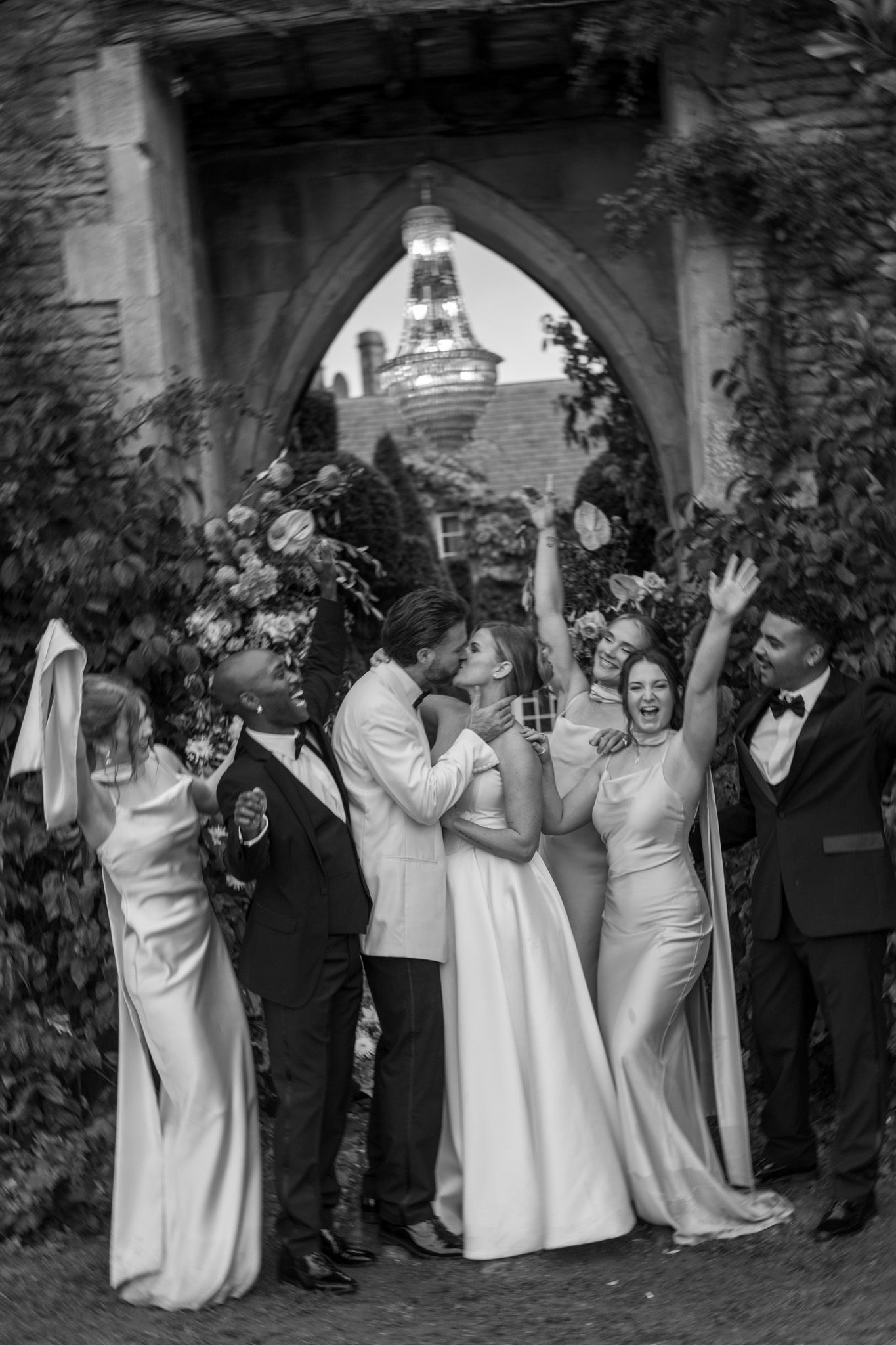 Black and white photo of wedding party celebrating under stone archway.