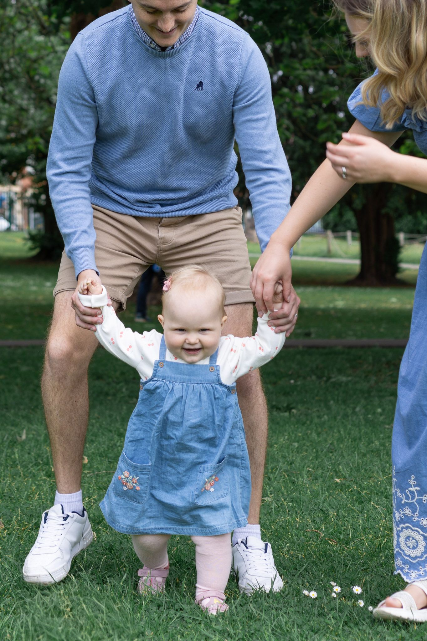 Baby in blue dress takes first steps with parents' help on green lawn.