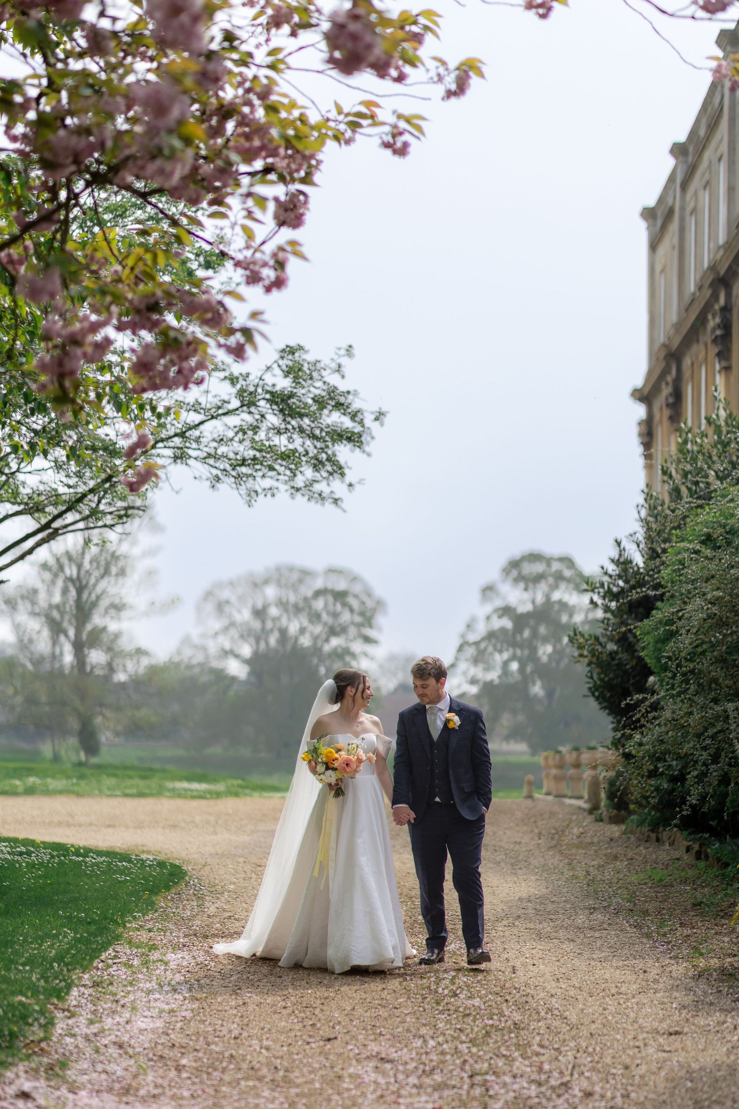 A couple walks along a gravel path under blooming cherry blossom trees on their wedding day.