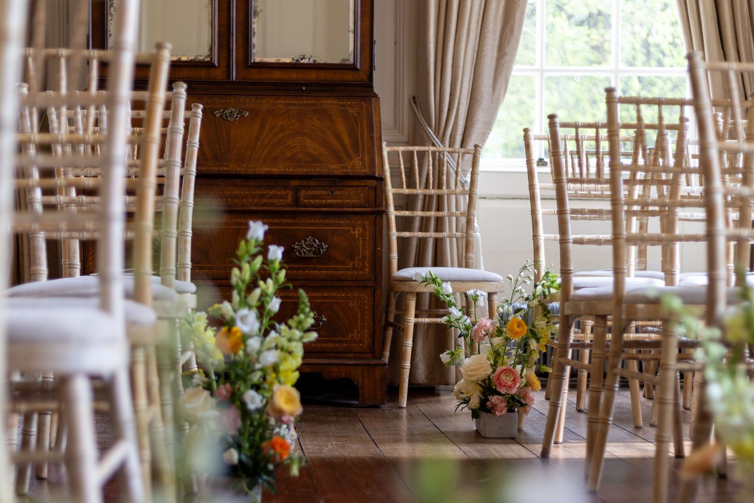 Elegant wedding ceremony setup with chiavari chairs lining an aisle adorned with colorful floral arrangements in a bright room.