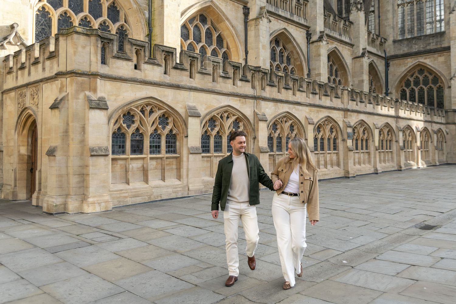 Couple holding hands walking past Bath Abbey's Gothic architecture on a sunny day.