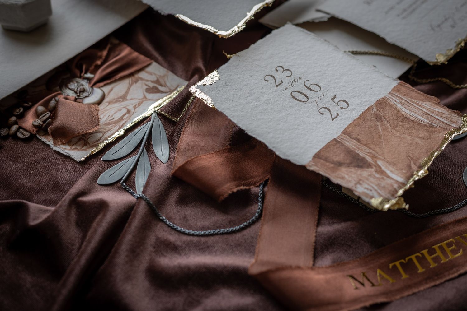 Brown satin wedding stationery flatlay with gold-edged invitation cards dated 23.06.25 and floral details.