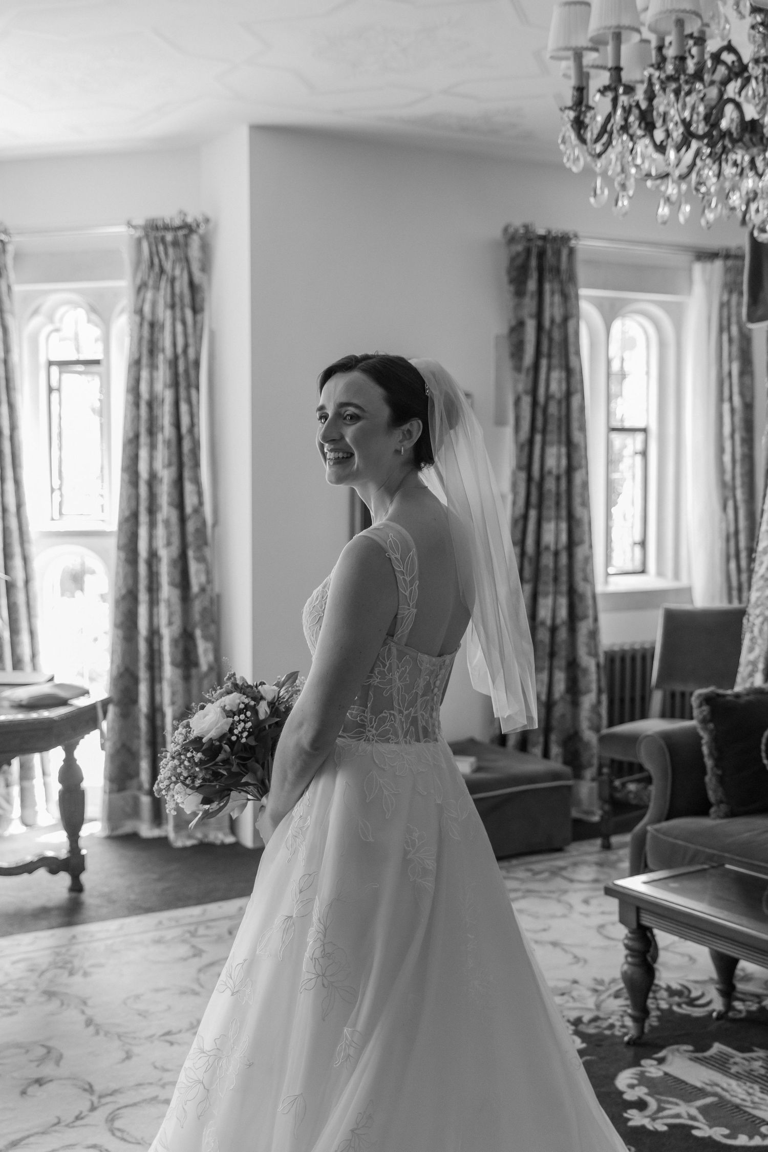 A smiling bride in a white lace gown and veil holds a bouquet in an elegant room with chandelier.