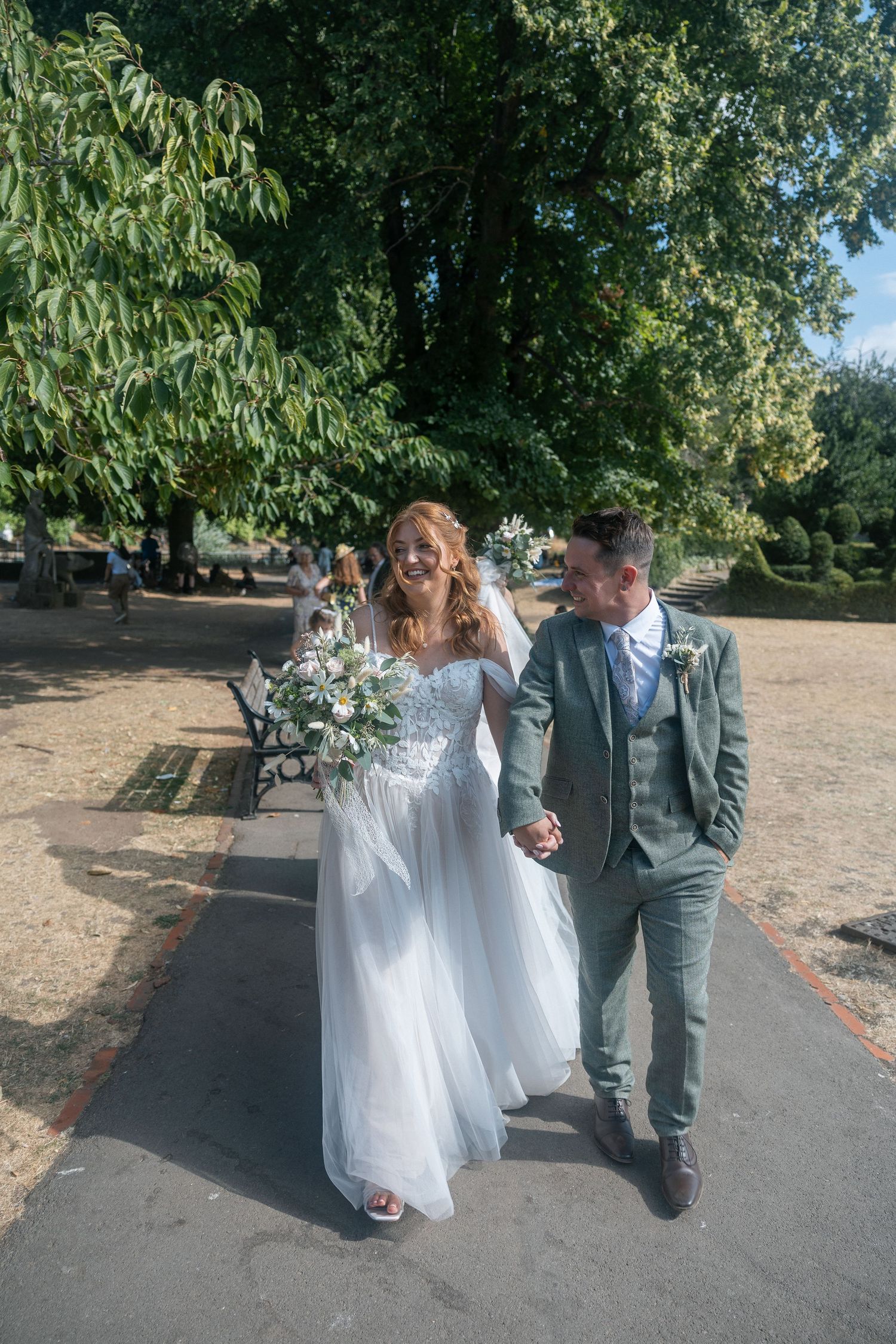 A bride and groom walk hand-in-hand through a sunny park, she in a white gown holding a bouquet, he in a sage suit.