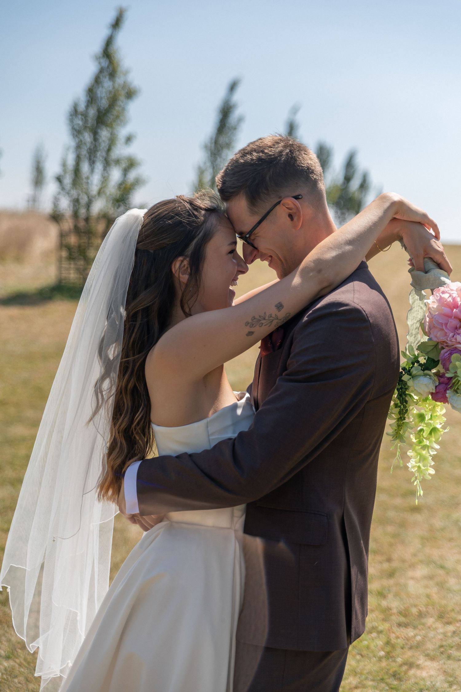 Bride and groom embrace outdoor Cripps Barn on sunny day, she holds pink bouquet wearing veil, he wears dark suit.