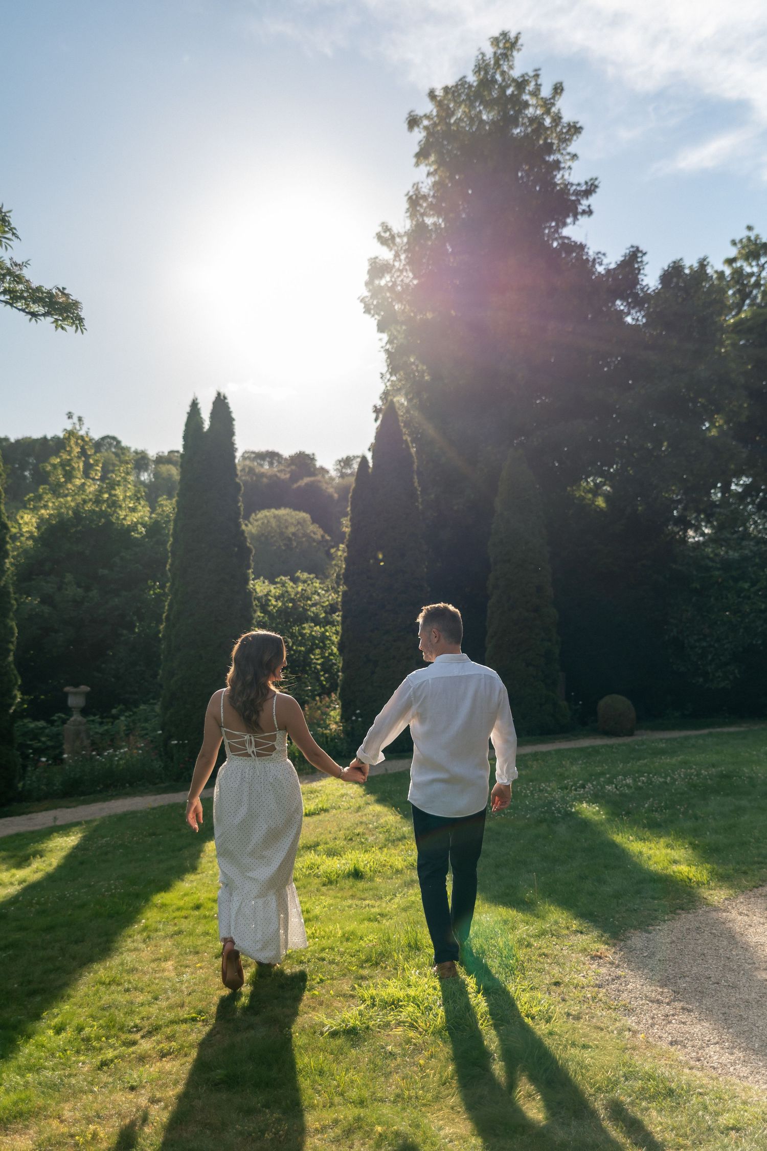 Couple holding hands walking through a sunlit garden with tall cypress trees and lush green lawn.