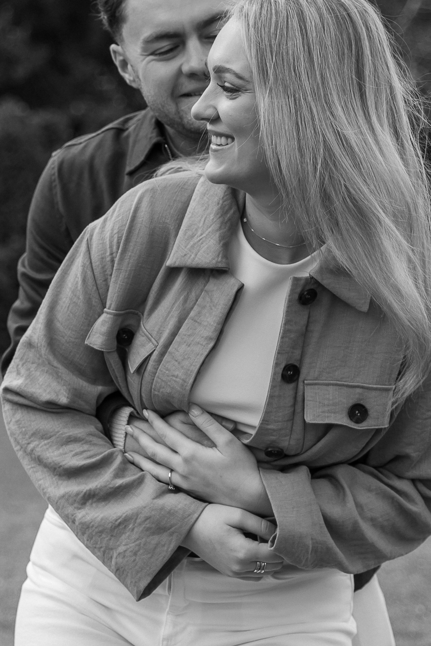 Black and white photo of a smiling couple embracing outdoors, woman in jacket and white pants, man hugging her from behind.