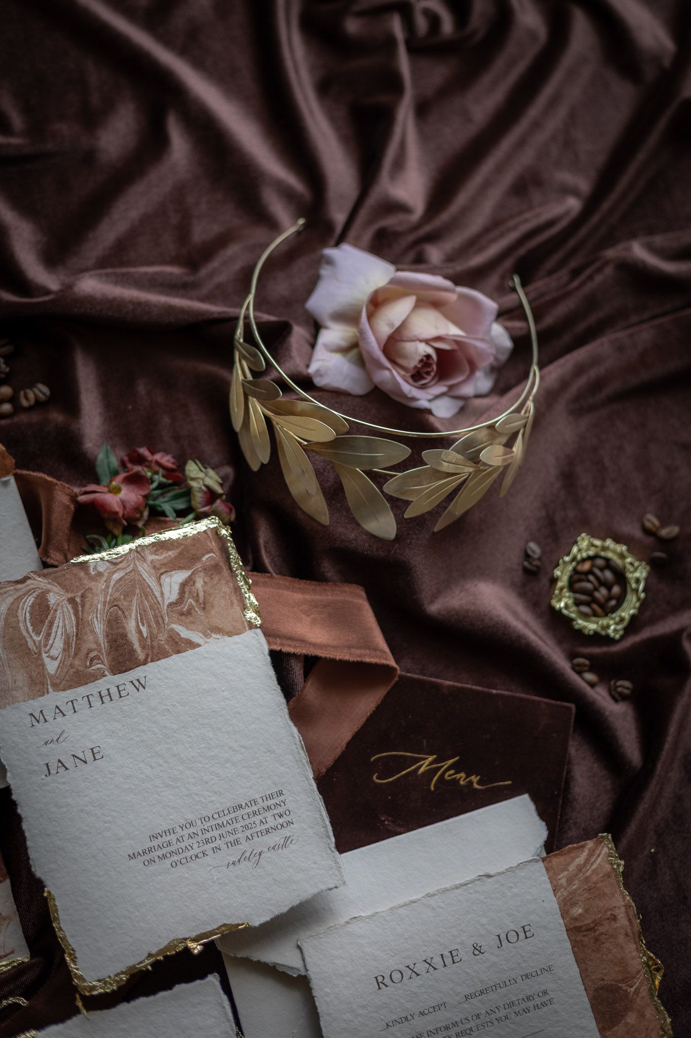Elegant wedding stationery flat lay with gold leaf crown, pink rose, and brown velvet backdrop featuring Matthew and Jane invite.