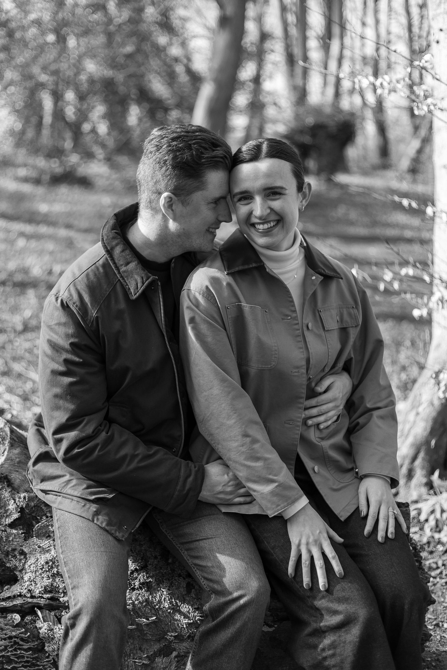 A couple shares an intimate moment while sitting together outdoors in a wooded setting, captured in black and white.