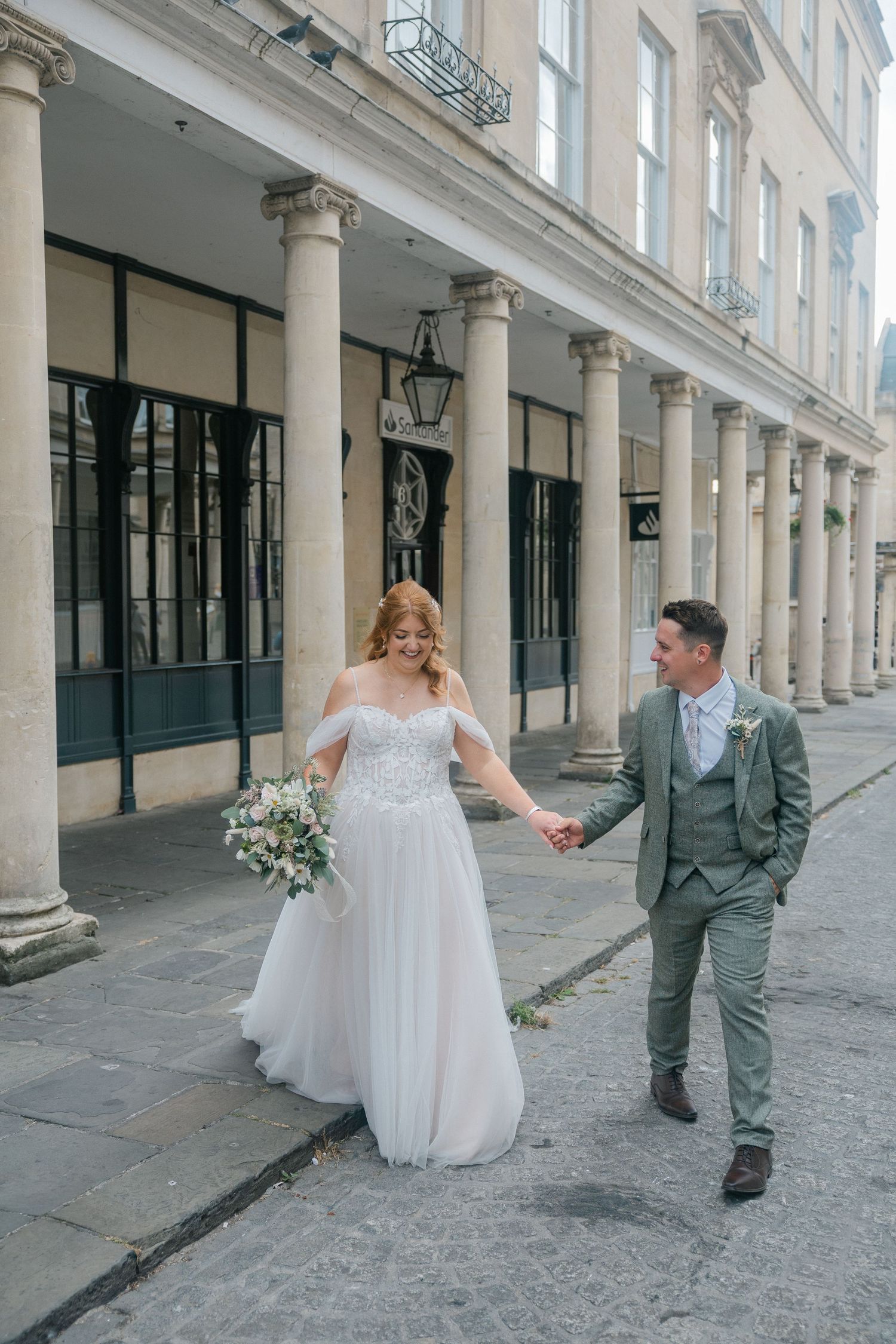 A bride and groom walk hand in hand outside a Georgian building with tall columns, she holds a bouquet.