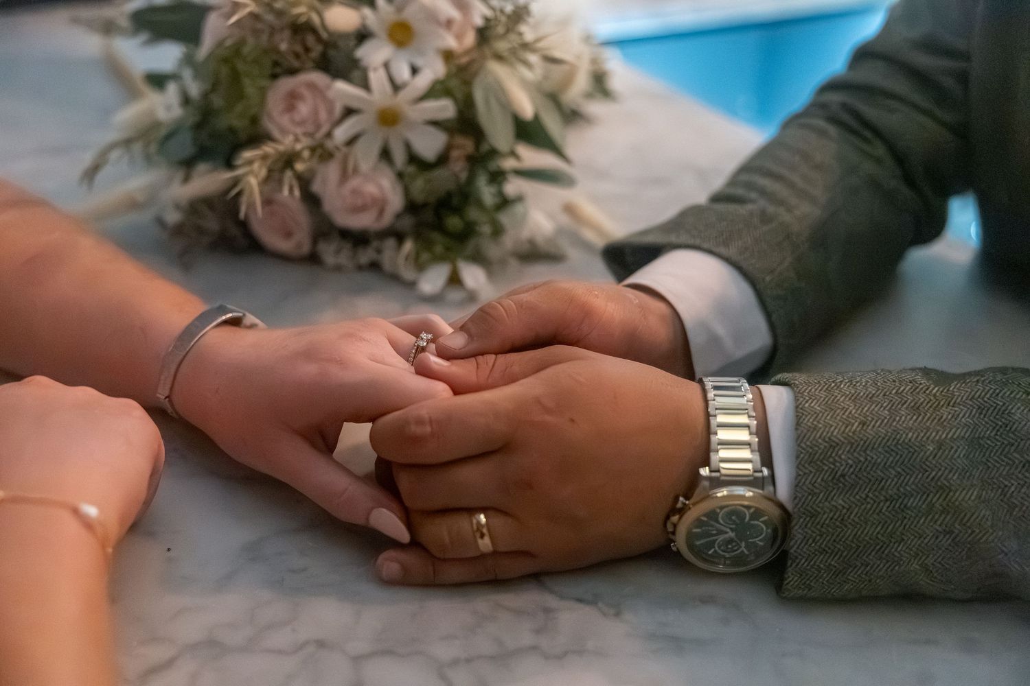 Couple holding hands with wedding rings on marble surface, bridal bouquet of roses in background.