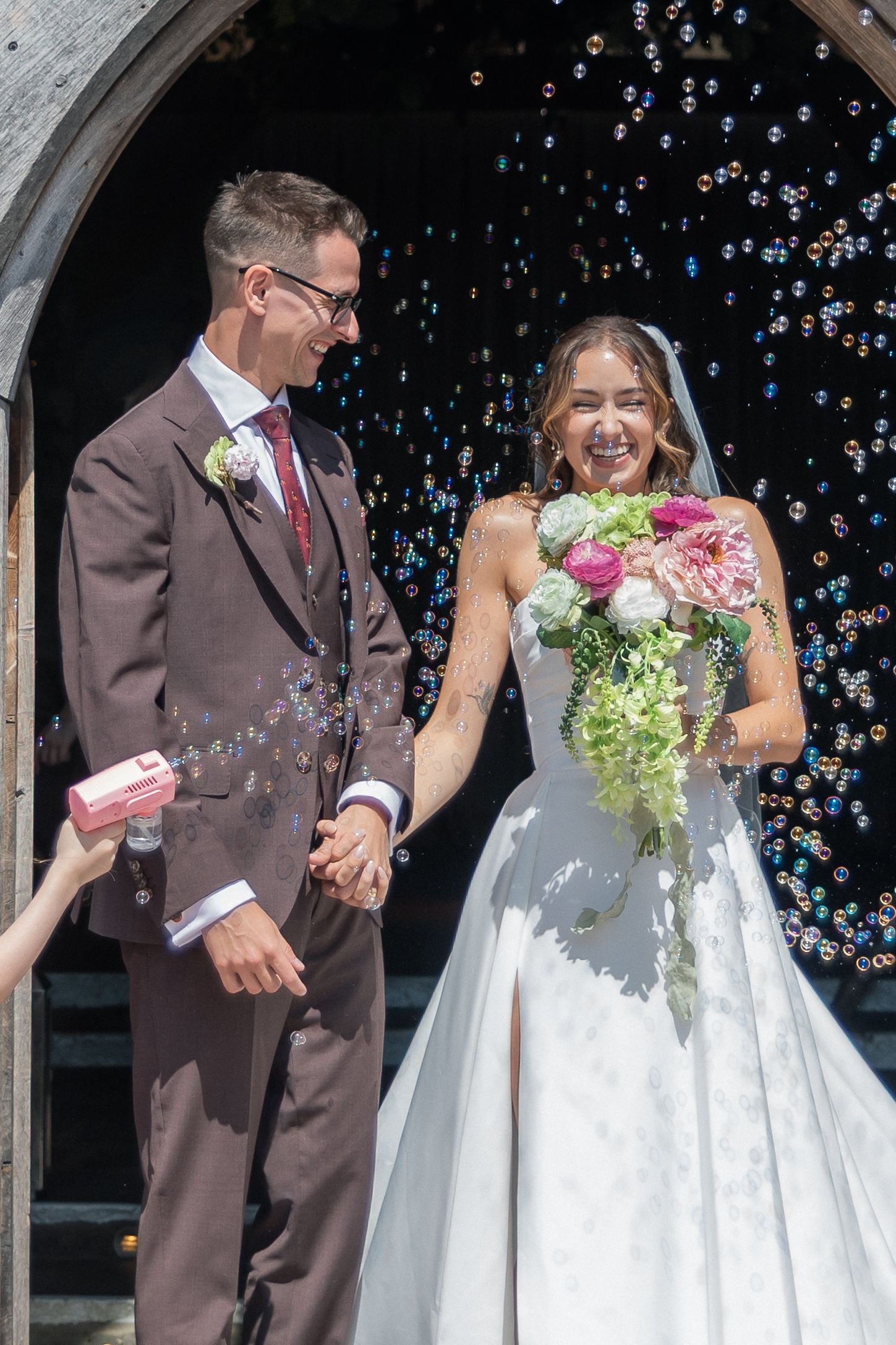 Joyful newlyweds exit church surrounded by bubbles, bride in white gown holding pink bouquet, groom in brown suit.