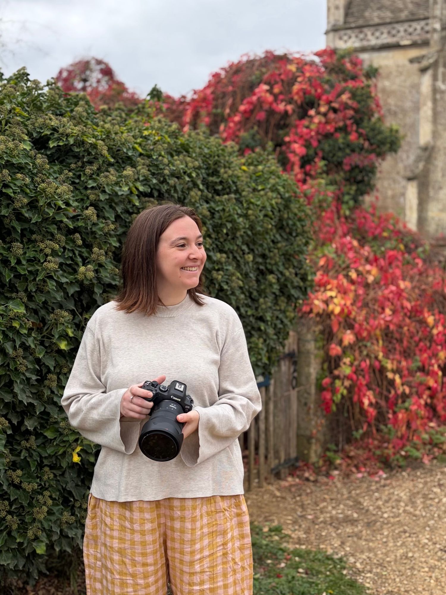 Woman holding a DSLR camera smiling outdoors with vibrant red autumn ivy and green hedges in background.