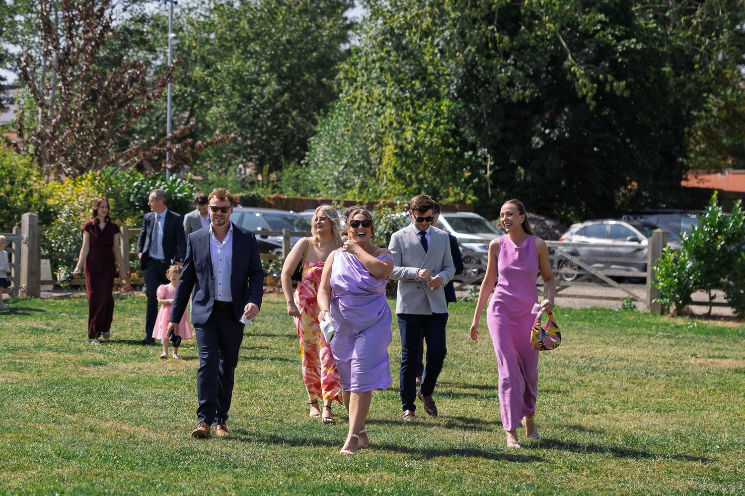 Wedding party walks together across a grassy lawn during an outdoor celebration on a sunny day.