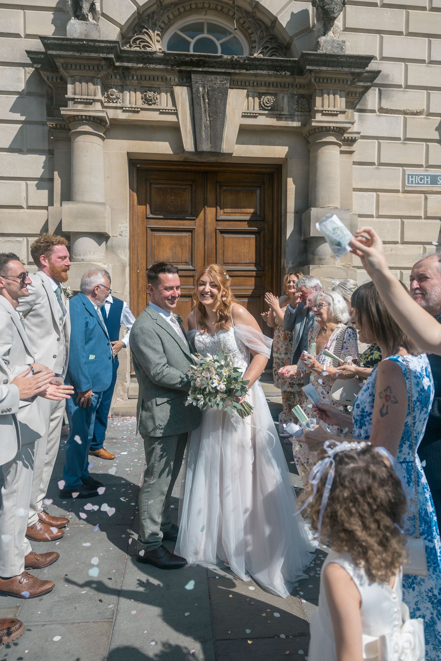 Bride and groom exit church as guests throw white confetti during sunny outdoor wedding ceremony celebration.
