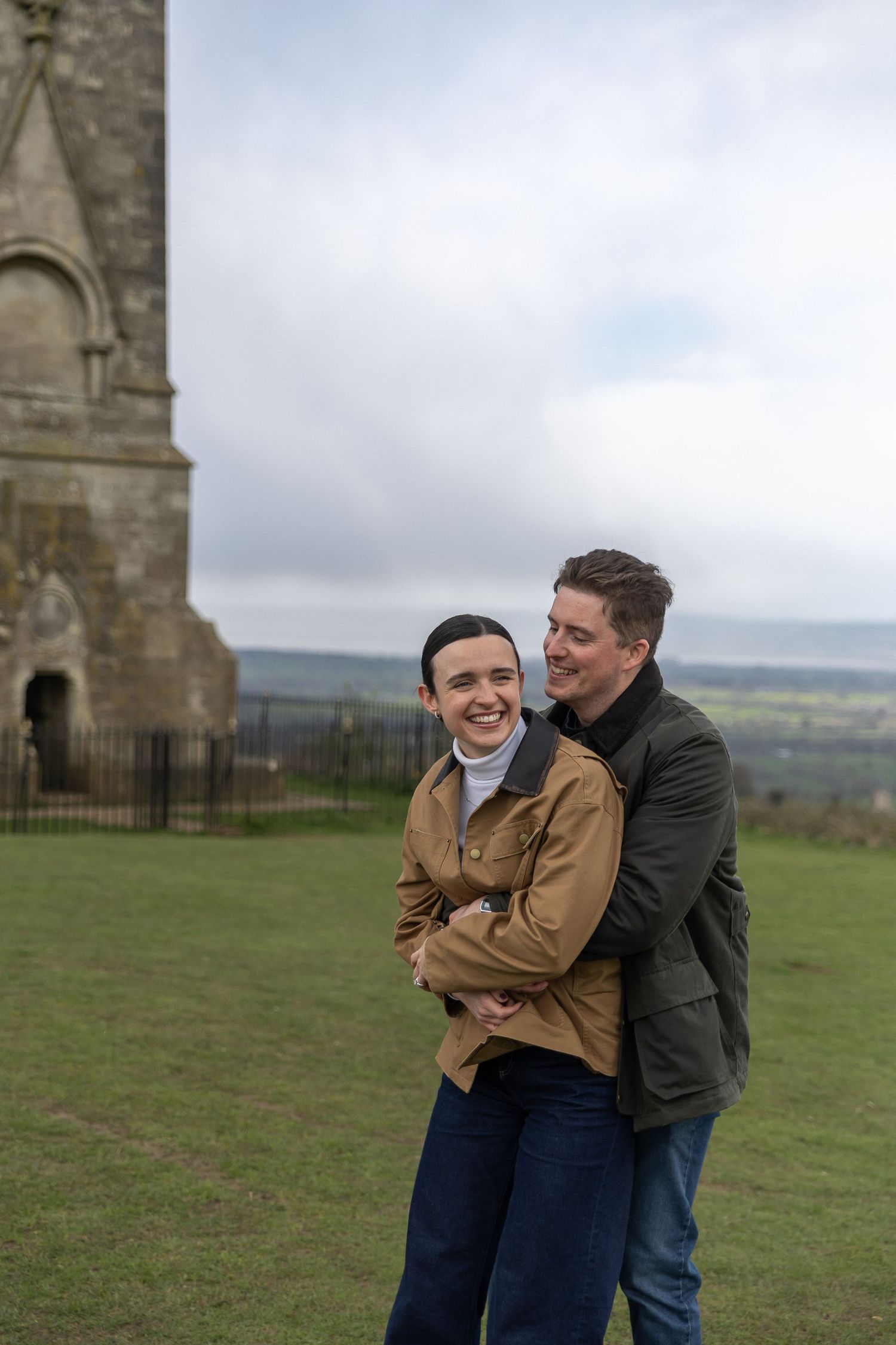A couple embraces and laughs together on a grassy hilltop with stone architecture visible in the background.