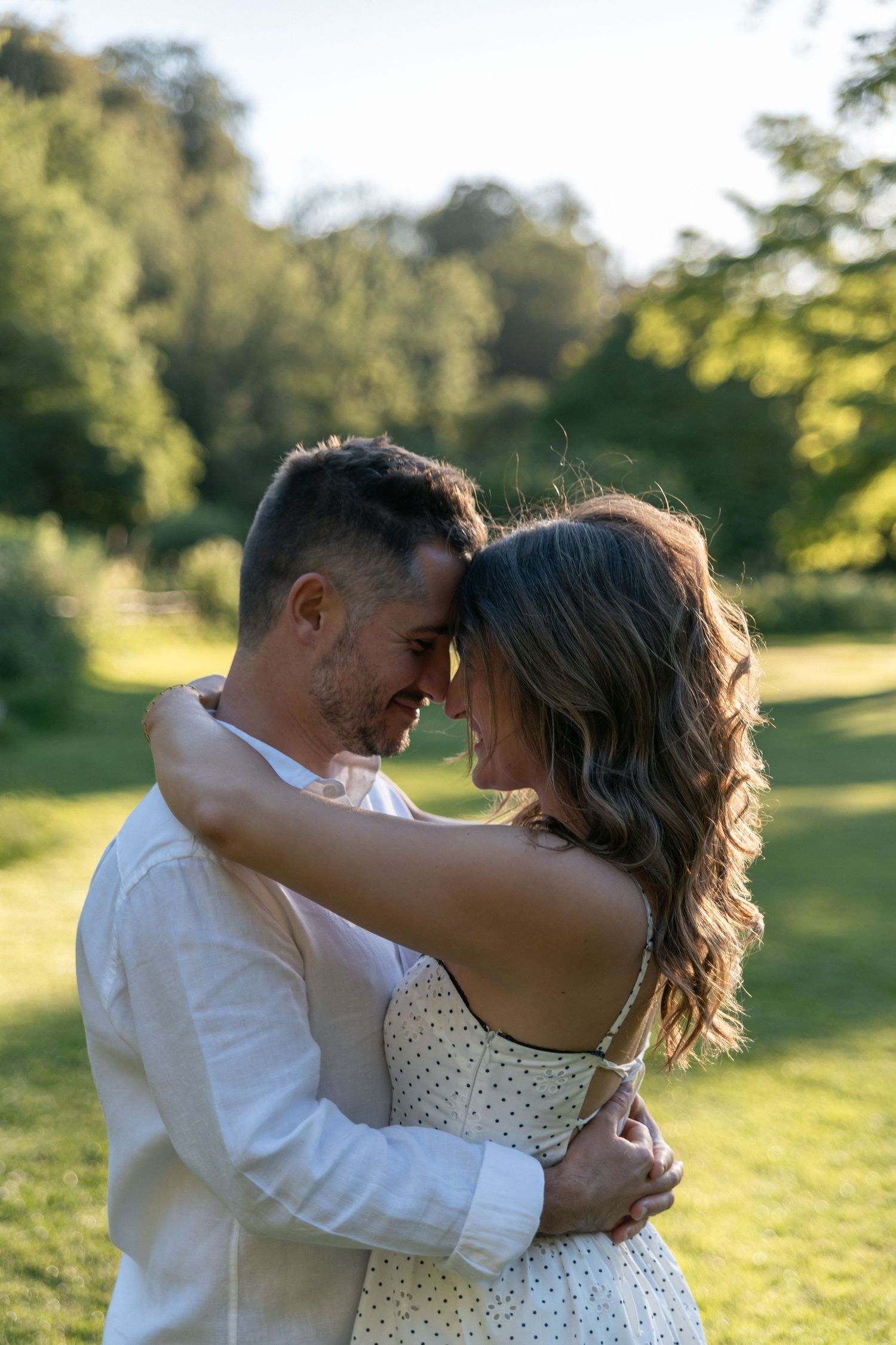 Couple embracing and smiling at each other in a sunny green park, wearing white outfits on a warm summer day.