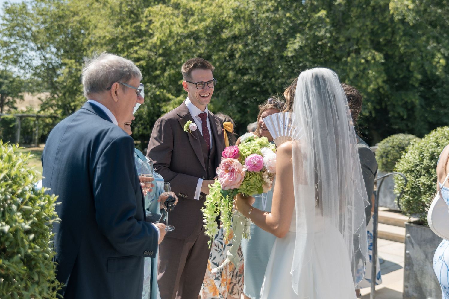 Groom in brown suit smiles at bride holding pink bouquet during outdoor wedding ceremony on sunny day.