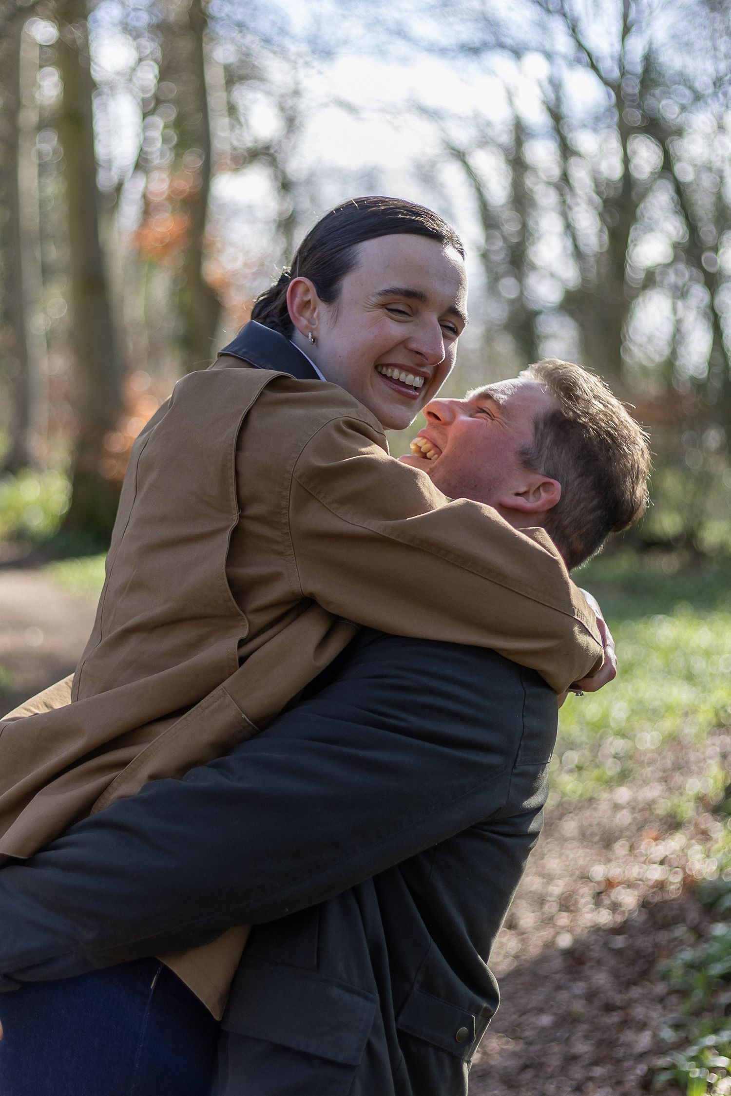 Two people share a joyful embrace outdoors in a wooded area on a sunny day.