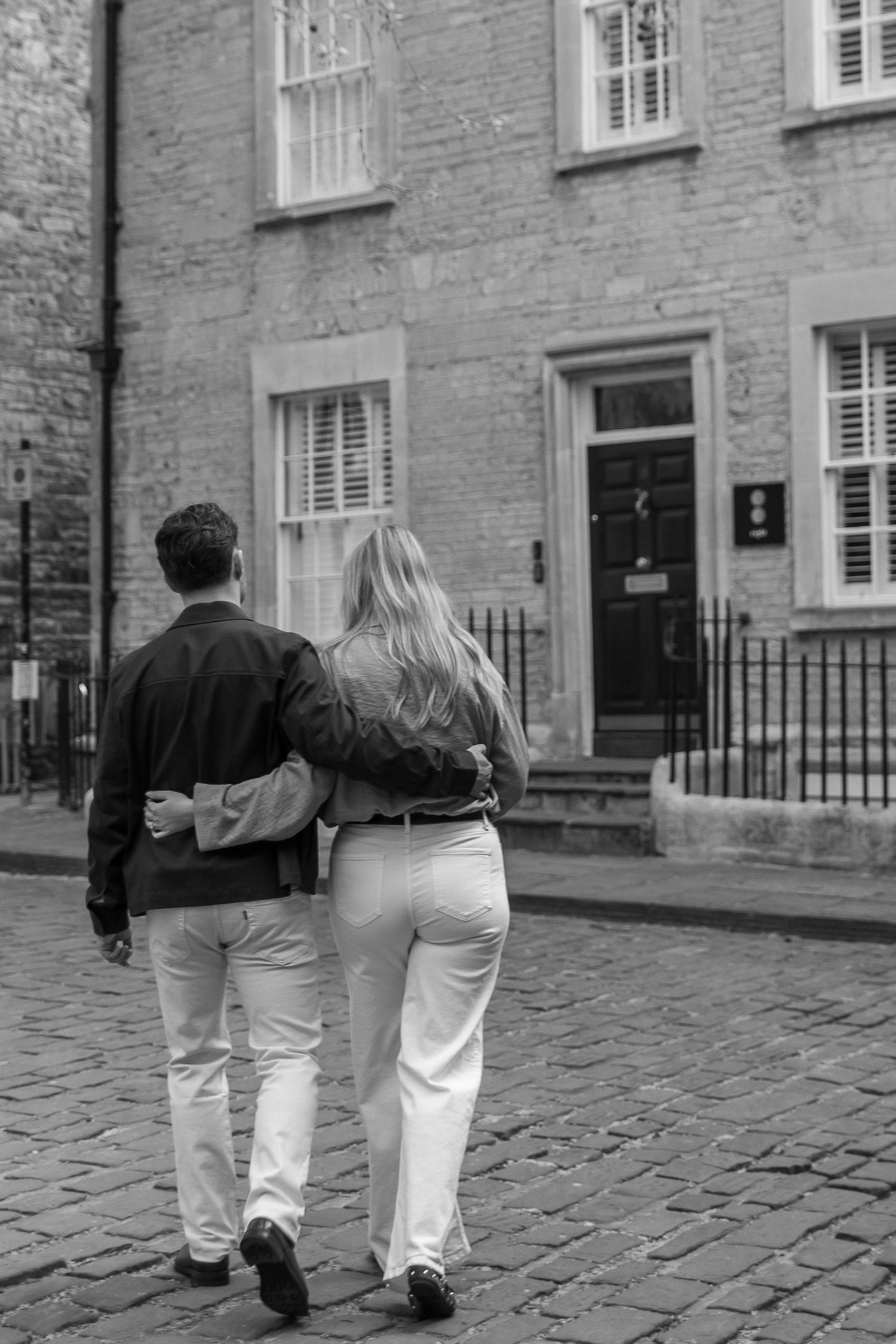 Couple walking arm in arm on cobblestone street past a classic brick building, black and white photo.