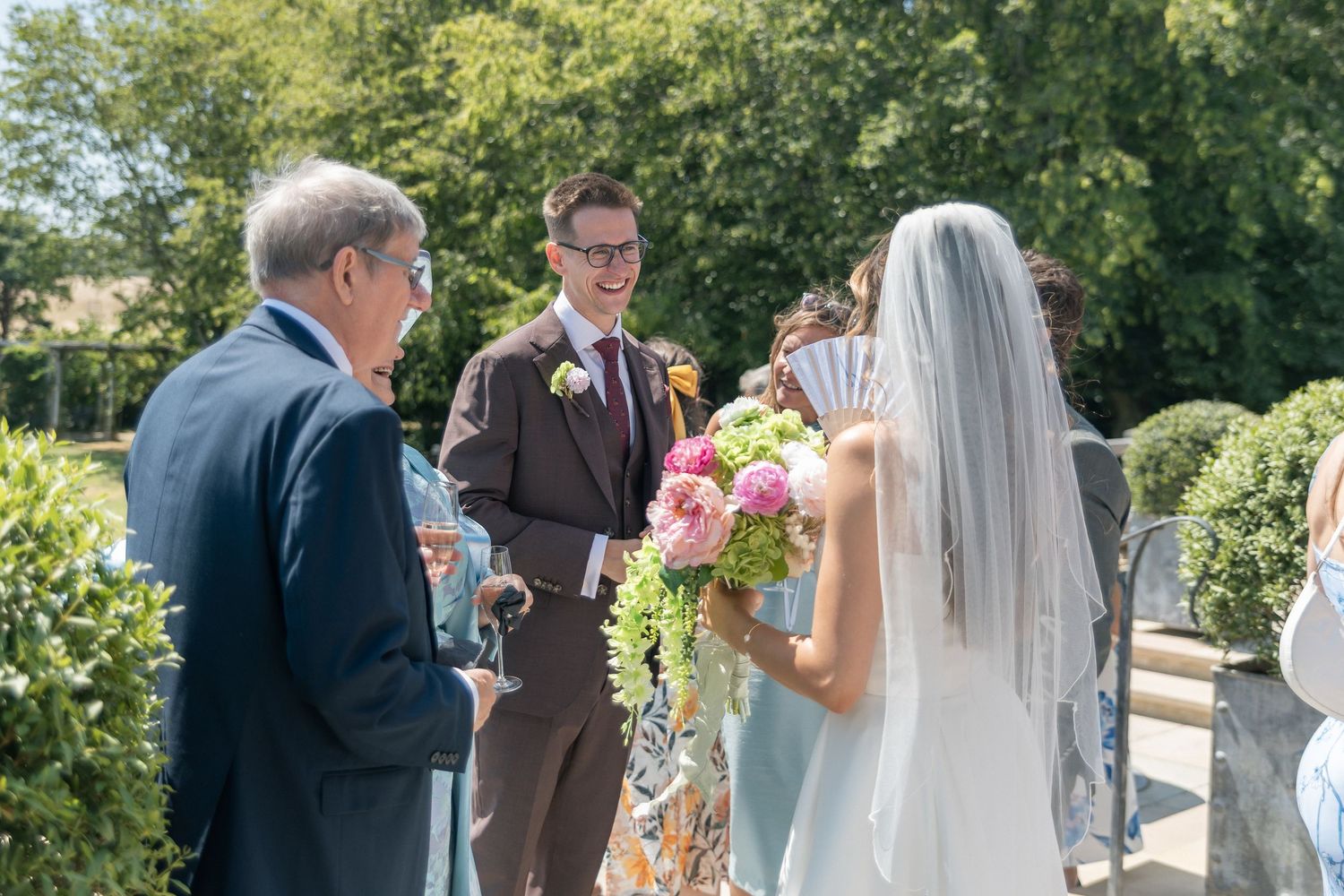 Groom in brown suit smiles at bride holding pink bouquet during outdoor wedding ceremony on sunny day.
