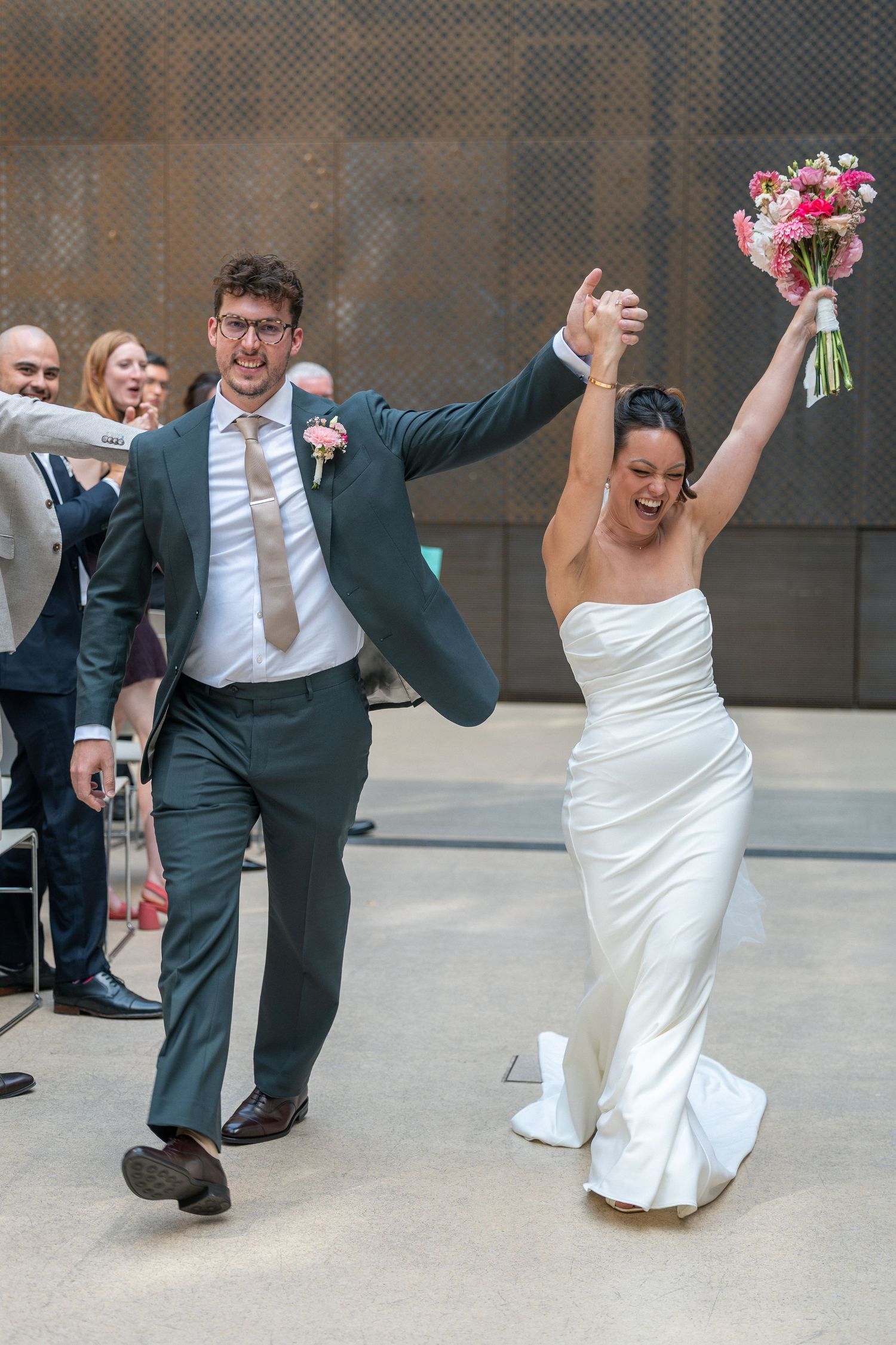 Joyful newlyweds celebrate after wedding ceremony, bride in white gown holding pink bouquet, groom in teal suit, guests cheering.