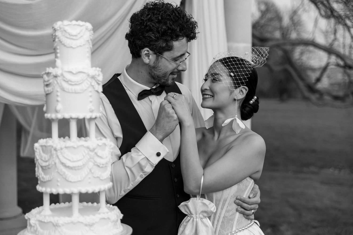 Black and white photo of a bride and groom smiling together beside a tall tiered wedding cake outdoors.