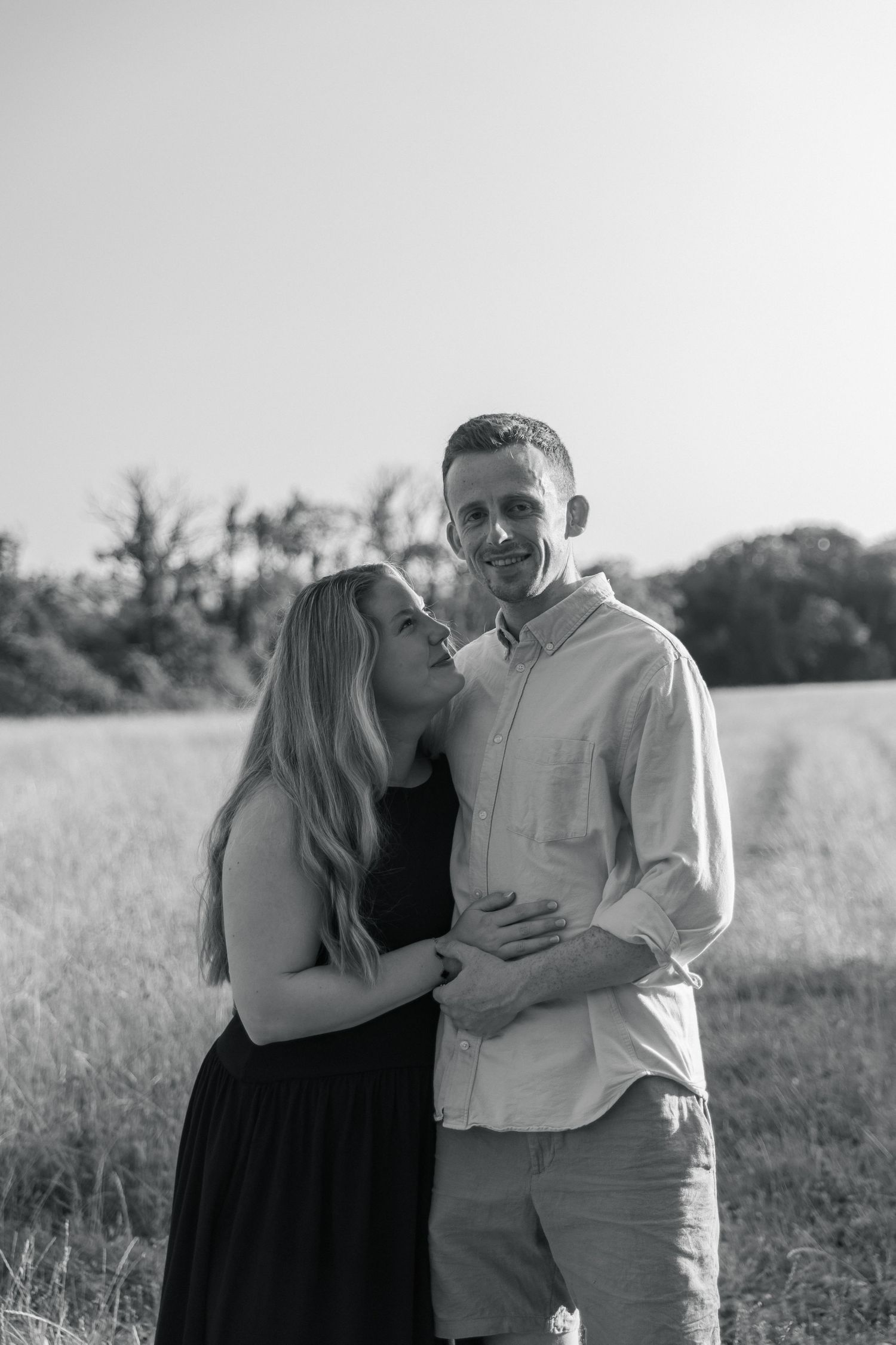 Black and white photo of a couple embracing in a field, woman kissing mans cheek while he smiles at camera.