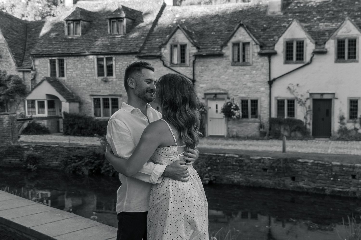 Black and white photo of a couple embracing and smiling by a river with charming stone cottages in the background.