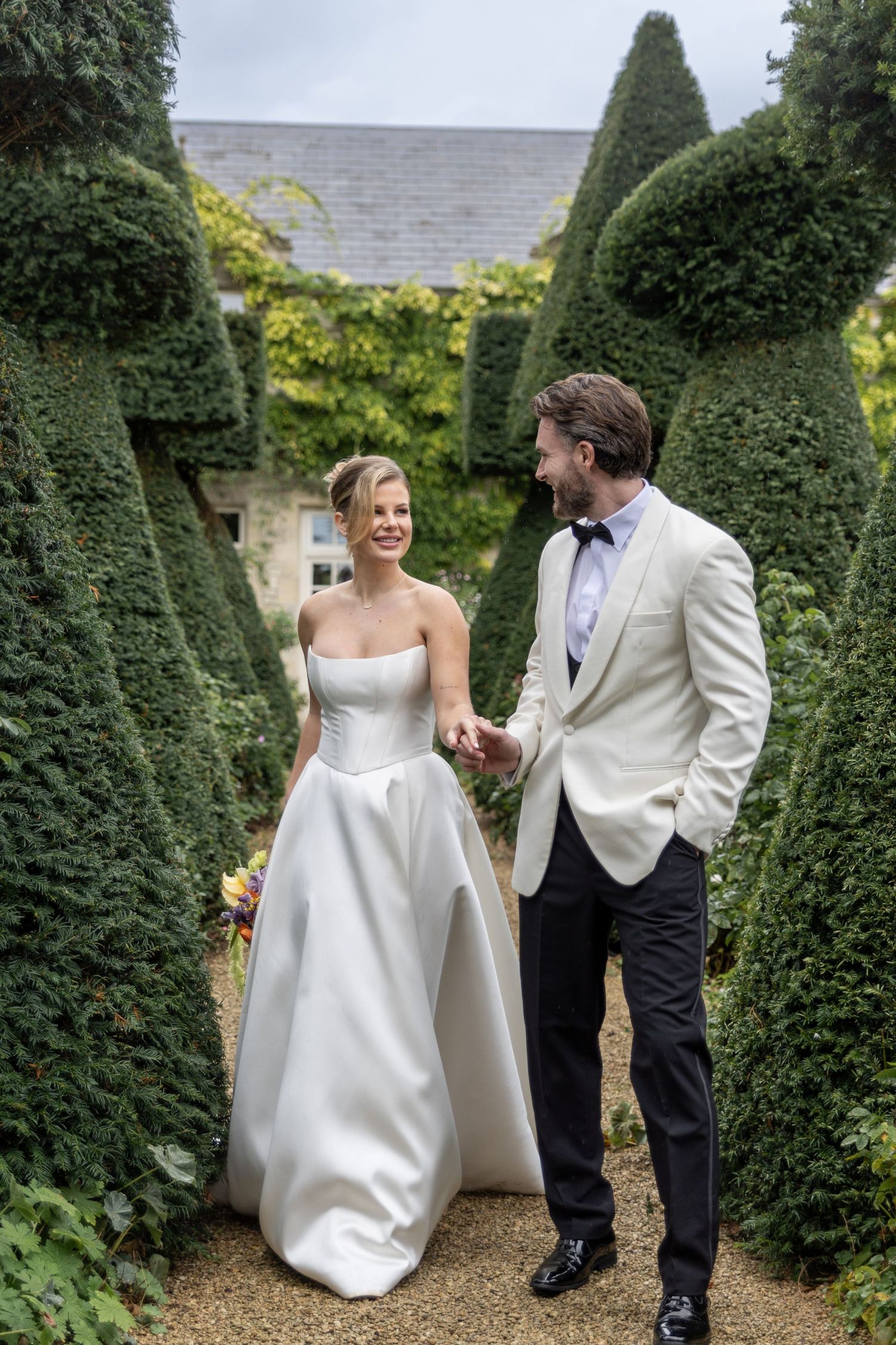 Newlywed couple shares joyful moments between manicured topiary bushes in an elegant garden setting.