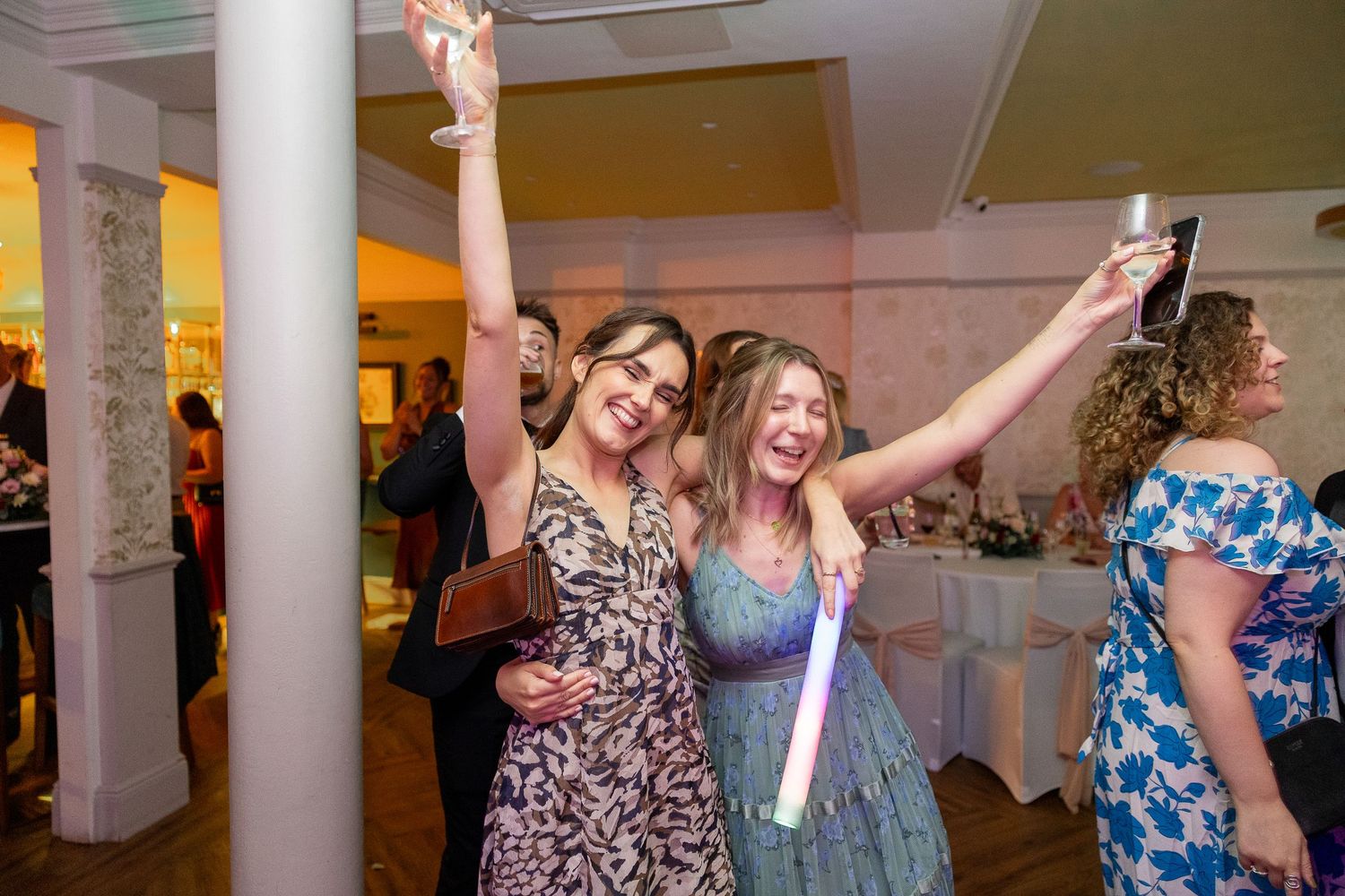 Wedding guests dancing and celebrating on a dance floor at a reception venue with white columns and warm lighting.