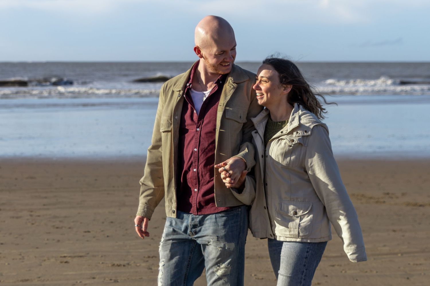 A couple walks hand in hand along a scenic beach shoreline on a sunny afternoon.