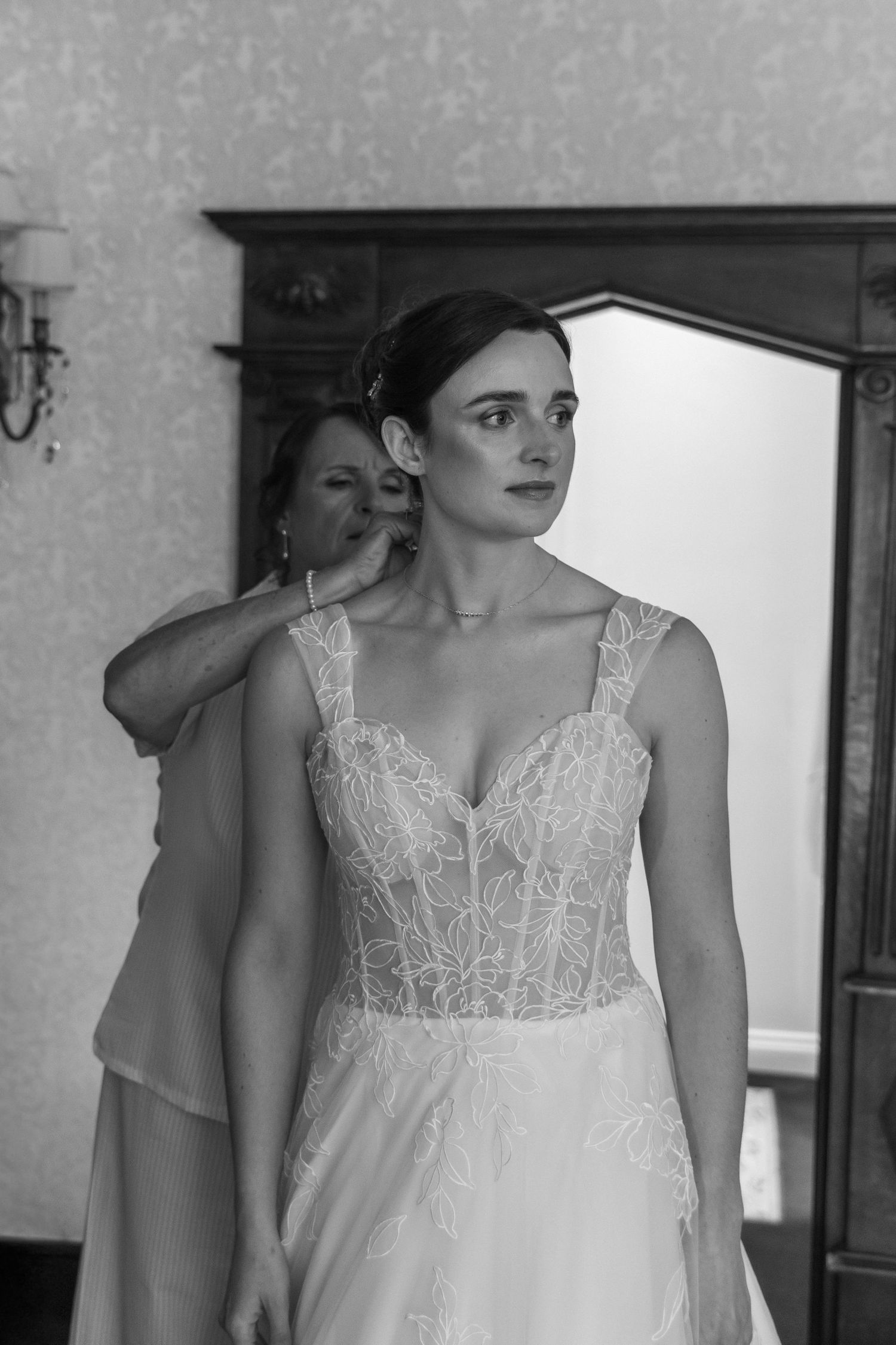 Black and white photo of a bride in lace wedding gown being helped with earring by bridesmaid in elegant room.